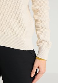 Sweter wełniany w kolorze kremowym z teksturowanym wzorem diamentowym, z prążkowanymi mankietami i dołem. Widać akcent w kolorze granatowego pomarańczu na mankiecie.