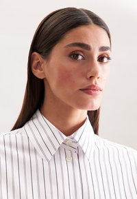 Jonge vrouw met lang, donker haar die een witte blouse met dunne verticale zwarte strepen draagt, peinzend naar de zijkant kijkend.