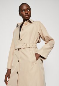Cappotto trench beige con vita regolabile, chiusura a bottoni e tasche laterali; presenta un colletto classico e un tessuto liscio e resistente all'acqua.