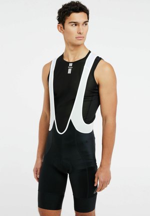 Jonge man in zwarte wielerbroek met bretels en mouwloos shirt, staand en naar de zijkant kijkend tegen een witte achtergrond.