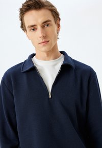 Marineblauw geribbeld sweater met een rits aan de voorkant en een gestructureerde kraag, gedragen over een wit T-shirt met ronde hals, gemaakt van een textuurstof.