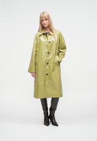 Grønn, blank, knelang regnfrakk med rett passform, stor krage og tre knapper. Brukes med svarte, knelange støvler.