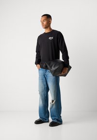 Junger Mann in einem schwarzen Sweatshirt und zerrissenen blauen Jeans, der eine große schwarze Lederhülle hält und vor einem schlichten weißen Hintergrund steht.