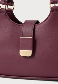 Burgundi värvi käekott tekstureeritud pinnaga, millel on kuldne pandlaaksent ja struktureeritud, pooleks ringikujuline kuju. Kaasas reguleeritav rihm.