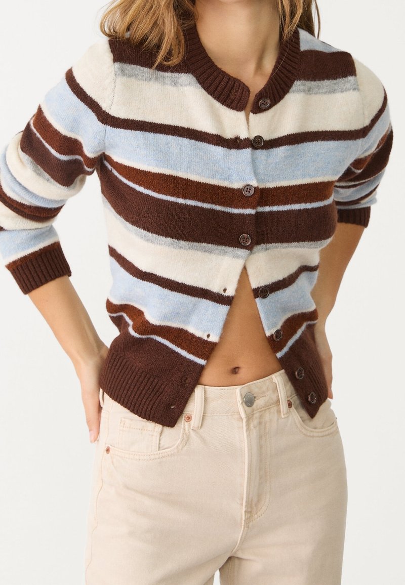 Femme portant un cardigan rayé boutonné marron, blanc, bleu clair et gris avec un pantalon taille haute couleur crème, mains posées sur les hanches.