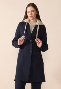 Cappotto navy con design su misura, caratterizzato da un cappuccio beige, due bottoni, tasche laterali e una texture morbida.