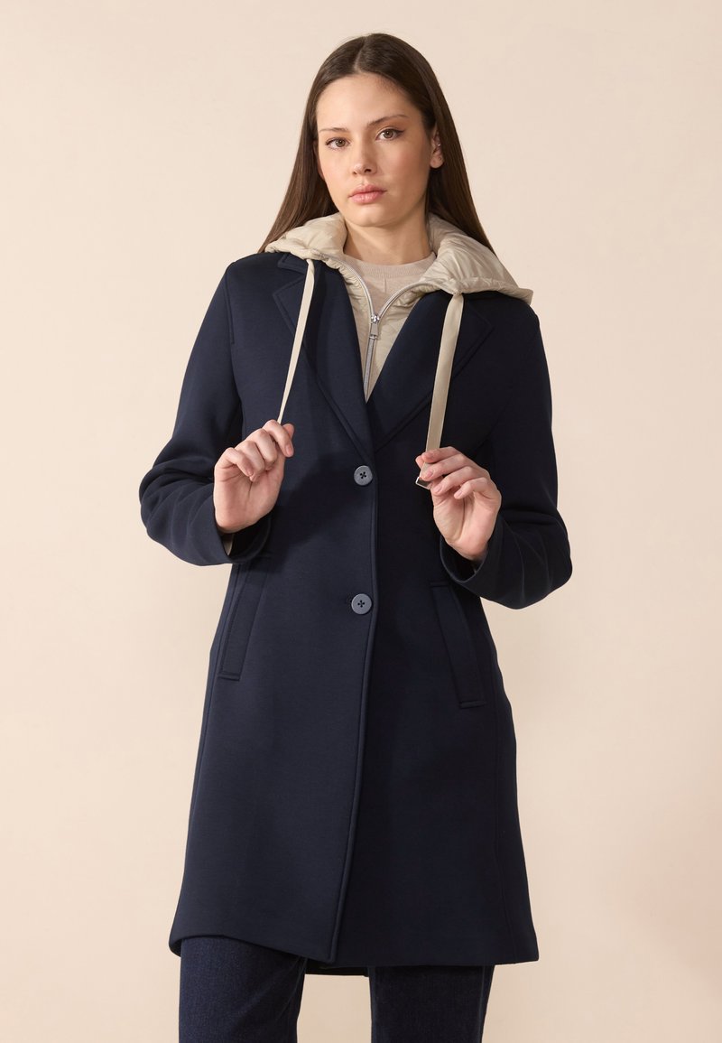 Cappotto navy con design su misura, caratterizzato da un cappuccio beige, due bottoni, tasche laterali e una texture morbida.