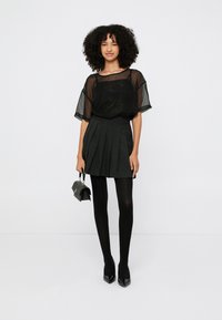 Schwarzes, transparentes Oberteil kombiniert mit einem plissierten schwarzen Rock, schwarzen Strumpfhosen und spitzen schwarzen Pumps. Kleine schwarze Handtasche als Accessoire.