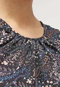 Černá květinová blůza se skládacím výstřihem; má malé bílé a světle modré květiny, složité vzory listů a jemnou texturu látky.