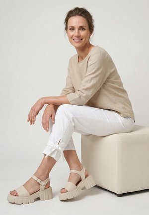 Frau mit braunen Haaren in einem beigen Langarmshirt und weißen Hosen mit Schleifen am Knöchel sitzt auf einem Würfelhocker und trägt beige klobige Sandalen.