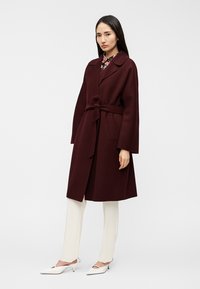 Μπορντό μάλλινο παλτό με ζώνη, γιακά με σκίσιμο και μακριά μανίκια, φορεμένο πάνω από λευκό παντελόνι και παπούτσια με τακούνι, σε ουδέτερο φόντο.