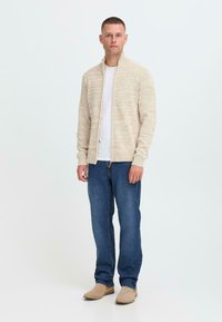 Mann som står, iført beige zip-up genser, hvit t-skjorte, blå jeans og beige semsket skinn støvler, mot en enkel hvit bakgrunn.