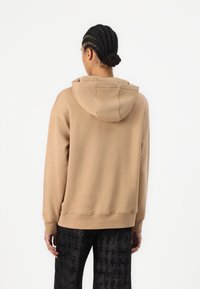 Hanorac bej cu glugă, cu o croială lejeră și manșete din material ribbed, prezentat din spate. Asortat cu pantaloni negri cu model. Textură fină.