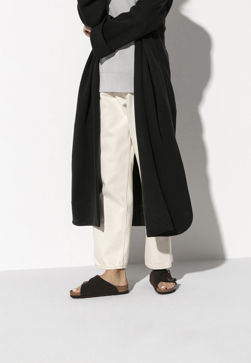 Un long manteau noir sur un haut gris, associé à un pantalon beige clair et des sandales à glissière marron foncé avec une semelle en liège. Les sandales ont un design ouvert.