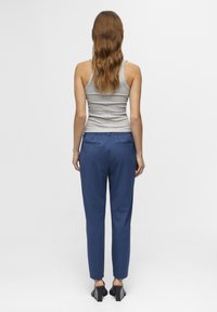 Een vrouw met lang, golvend haar staat met haar rug naar de camera, gekleed in een grijze tanktop en donkerblauwe broek, met hoge hakken sandalen.