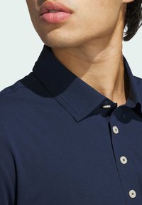 Granatowa polo z szerokim kołnierzykiem, trzema guzikami z jasnobeżowymi akcentami oraz gładką fakturą materiału. Zbliżenie na dekolt i kołnierzyk.