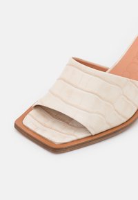Chaussure à enfiler de couleur crème avec un motif crocodile texturé et en relief, bout carré et semelle extérieure marron. Design minimaliste avec un dessus ouvert.