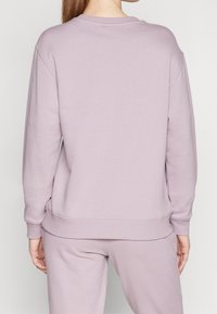 Person klädd i en enkel ljuslila sweatshirt och matchande byxor, sedd bakifrån mot en enfärgad bakgrund.