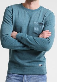 Maglione pullover teal realizzato in morbido tessuto, con una tasca sul petto a motivo, polsini a coste e un orlo dritto con un piccolo etichetta logo.