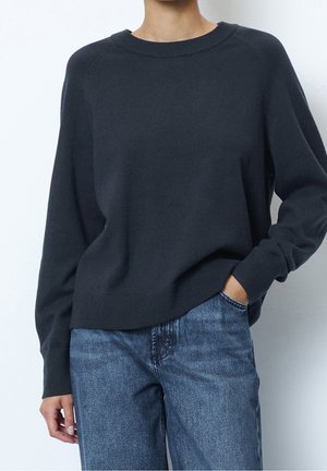 Nainen, jolla on päällään tummanharmaa pitkähihainen neule ja siniset farkut, toinen käsi taskussa yksinkertaista vaaleaa taustaa vasten.