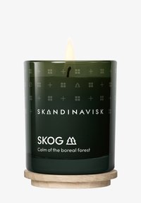 Green glass candle with a glowing flame. Textured surface featuring geometric patterns. Label reads “SKOG, Calm of the boreal forest.” Wood base.