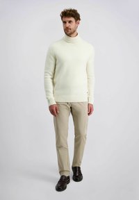 Crème turtleneck sweater met een geribde zoom en manchetten, gecombineerd met beige broeken en donkerbruine schoenen, staand op een neutrale achtergrond.