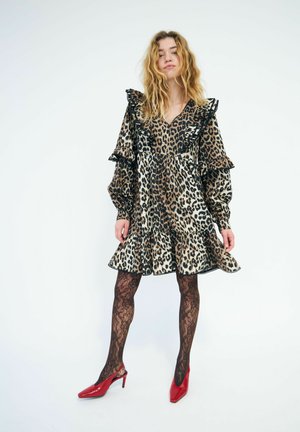 Femme aux cheveux bouclés porte une robe à volants imprimée léopard, des collants en dentelle noire et des chaussures à talons rouges, debout devant un fond blanc.