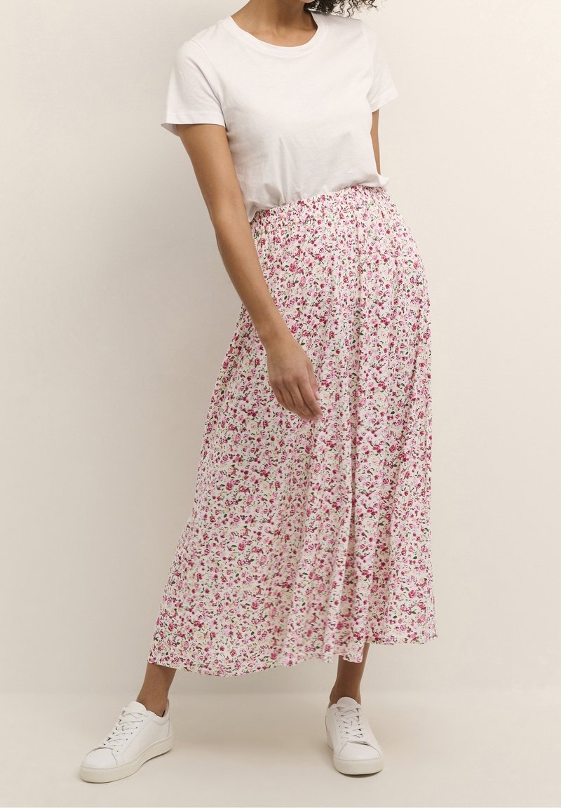 Femme portant un t-shirt blanc, des baskets blanches et une longue jupe blanche avec un petit motif floral rose, debout devant un fond uni.