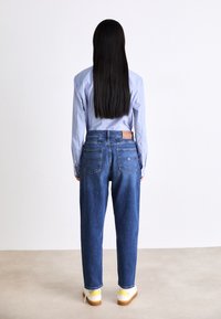 Vaqueros de denim azul con corte recto, cinco bolsillos y una etiqueta de cuero marrón. Combinados con una camiseta azul clara y unas zapatillas blancas/amarillas.