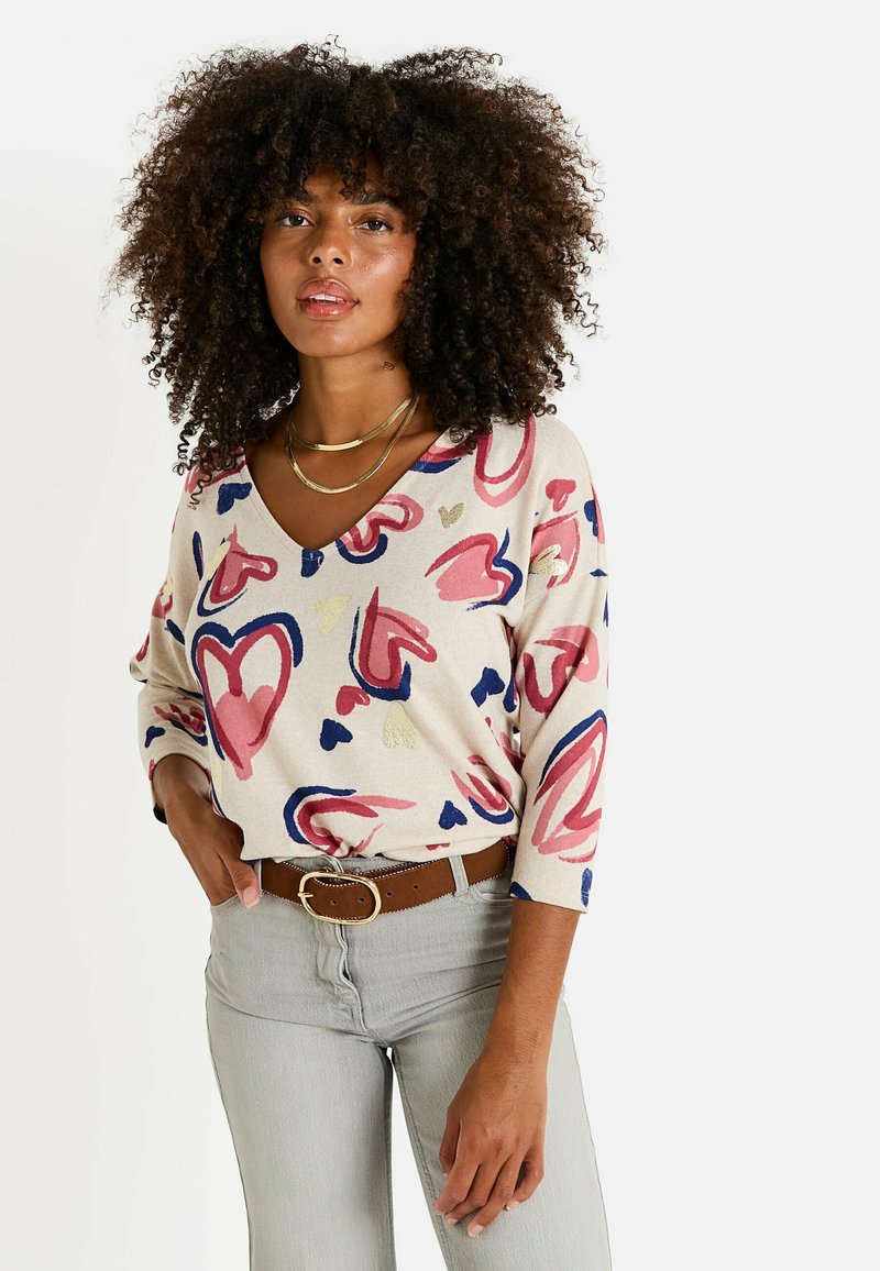Top à manches longues en V beige avec des motifs de coeurs colorés en rose, bleu et or. Associé à un jean gris et une ceinture marron.