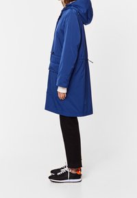 Blå regnjacka med huva, åtstramad midja och framfickor, parat med svarta byxor och svarta sneakers med röda detaljer.