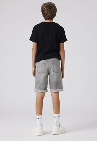 T-shirt noir, shorts en denim gris avec des revers, baskets blanches et chaussettes avec un logo. Tenue simple et décontractée vue de dos.