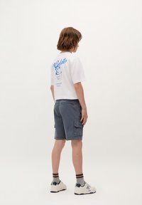 T-shirt bianco a maniche corte con stampa grafica blu sul retro, abbinato a pantaloni cargo grigi scuri e sneakers bianche con dettagli blu navy.