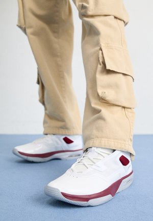 Beige cargobukser båret med hvide sneakers med mørkerøde accenter stående på et lyseblåt tæppe.