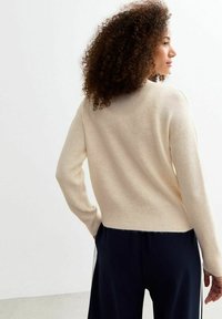 Maglione color crema realizzato in materiale morbido, con un design corto, maniche lunghe e orlo a coste. Indossato con pantaloni scuri.