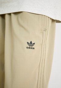 Beige Adidas φόρμες αθλητισμού διαθέτουν μαύρο λογότυπο και τρεις κάθετες ρίγες στο πλάι, κατασκευασμένες από λείο, ελαφρύ υλικό.