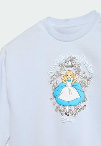 Ανοιχτό μπλε πουλόβερ με την Αλίκη σε λευκό και μπλε φόρεμα, περιτριγυρισμένη από λευκά μαργαρίτες και το κείμενο «Ο περίεργος κήπος της Αλίκης». Λογότυπο της Disney από κάτω.