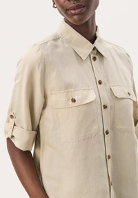 Camicia beige a maniche corte con bottoni, due tasche sul petto, maniche arrotolate e bottoni marroni. Tessuto morbido con una vestibilità comoda.