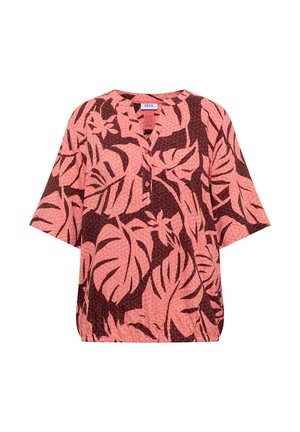 Femme aux cheveux courts et bruns portant une chemise ample rose et rouge foncé à motifs de feuilles tropicales et un pantalon beige, vue de dos.