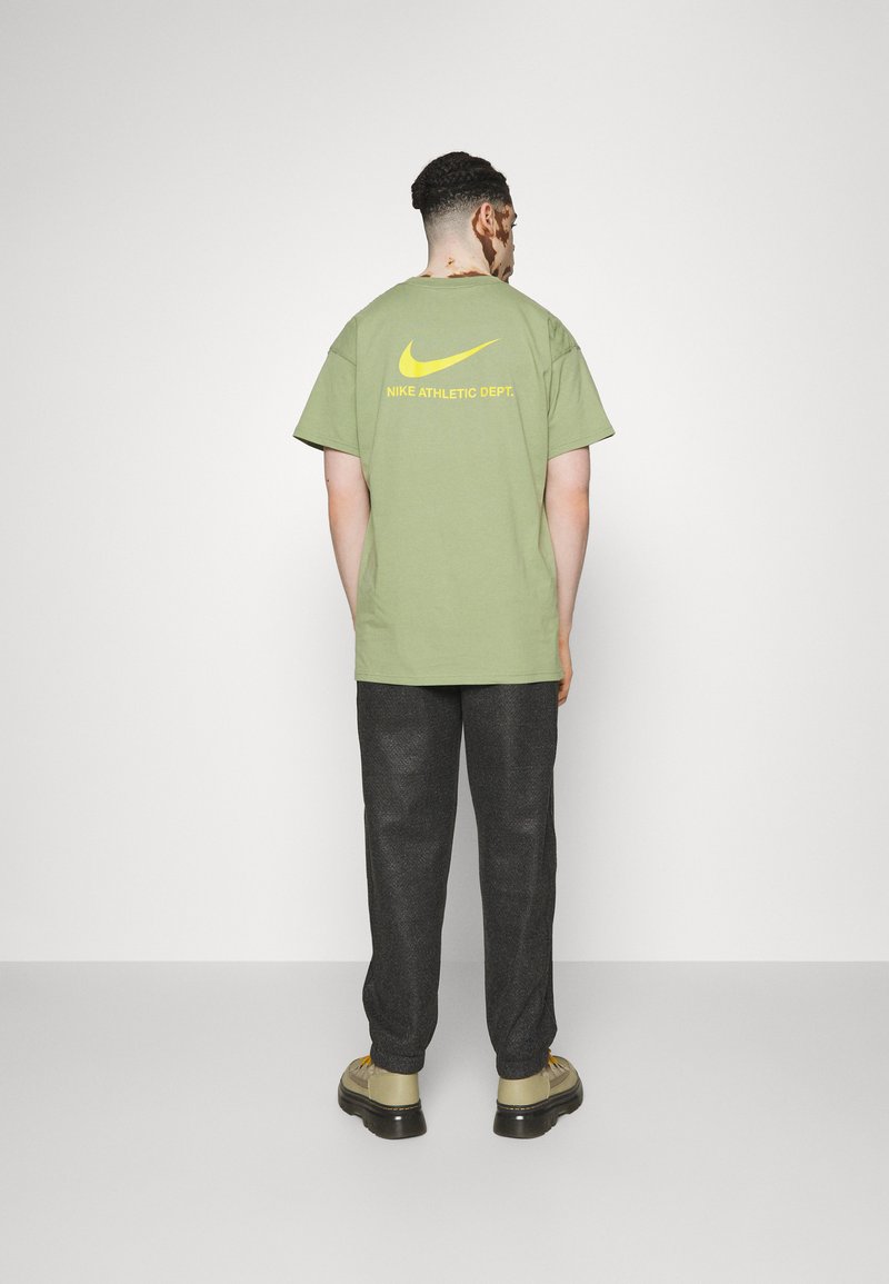 T-shirt en coton vert avec le logo Nike jaune et texte au dos, associé à un pantalon texturé sombre et des chaussures beige.