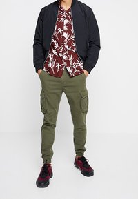 Uomo che indossa una giacca bomber nera, una camicia con stampa tropicale bordeaux, pantaloni cargo oliva e sneaker bordeaux e nere, in piedi con le mani in tasca.