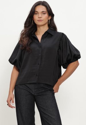 Woman wearing a black button-up blouse with voluminous puff sleeves and dark high-waisted pants, posing with one hand on her hip.