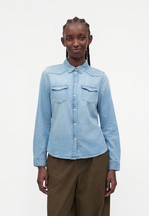 Kvinde med flettet hår iført en lyseblå denimskjorte med knapper og brune bukser, stående foran en ensfarvet hvid baggrund.