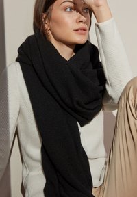 Sort tørklæde lavet af blødt stof, draperet om halsen, båret over en struktureret hvid sweater. Lyset rammer materialet og fremhæver dets tekstur.