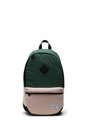 Sac à dos vert et beige de forme arrondie, fabriqué en tissu, avec une poche avant, une fermeture éclair noire et un écusson logo sur le bas de l'avant.