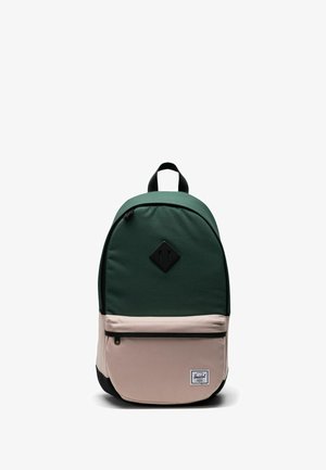 Sac à dos vert et beige de forme arrondie, fabriqué en tissu, avec une poche avant, une fermeture éclair noire et un écusson logo sur le bas de l'avant.