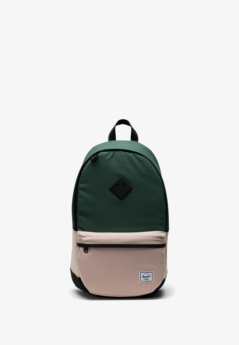 Sac à dos vert et beige de forme arrondie, fabriqué en tissu, avec une poche avant, une fermeture éclair noire et un écusson logo sur le bas de l'avant.