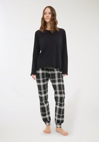 Giovane donna che indossa una maglia nera a maniche lunghe e pantaloni del pigiama a quadri bianchi e neri, in piedi a piedi nudi su uno sfondo semplice.