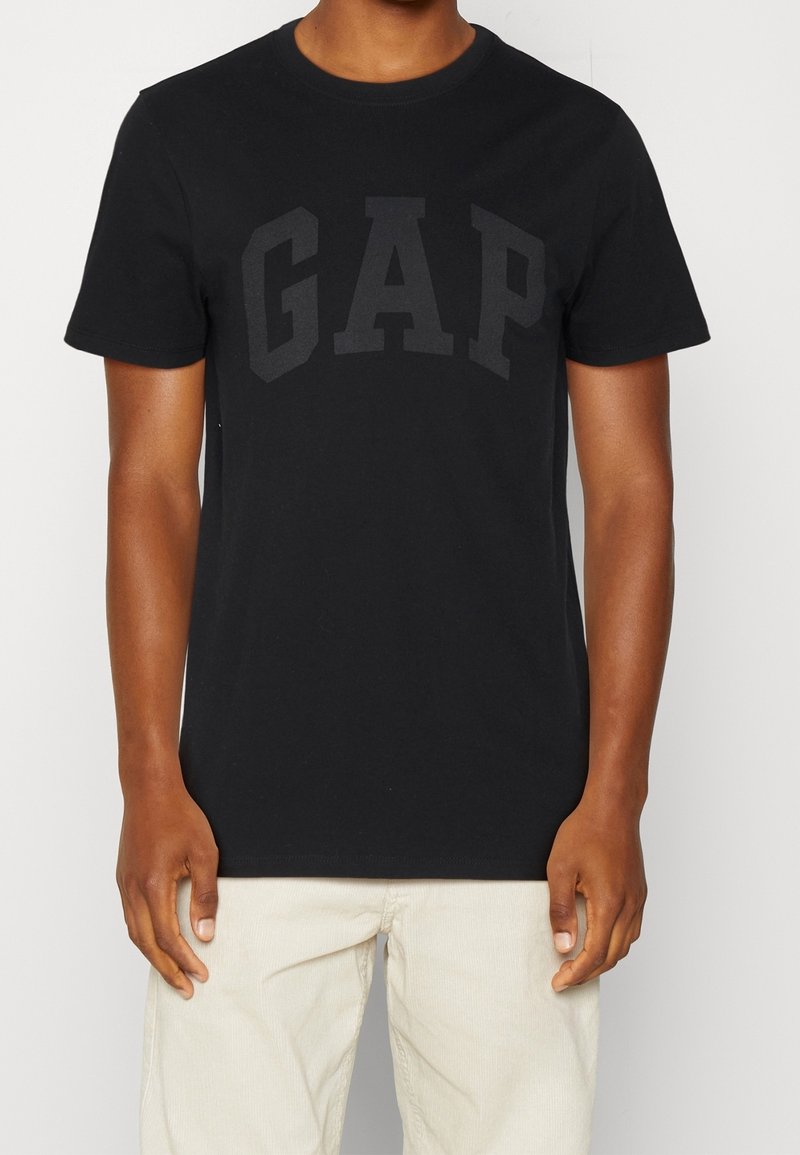 Homme portant un t-shirt noir à manches courtes avec un logo sombre "GAP" sur la poitrine et un pantalon clair, debout devant un fond blanc.