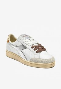 Sneaker bianco in pelle con lacci marroni, dettagli in argento texturizzato e suola in gomma beige. Presenta una morbida punta in camoscio e un tallone con un motivo unico.